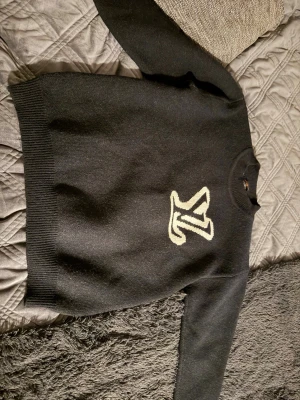 Svart stickad tröja  - Snygg svart stickad tröja med en stor vit logga på bröstet. Tröjan har rund halsringning, långa ärmar och ribbstickade muddar vid ärmslut och nederkant. Perfekt för dig som gillar stilrena plagg med en twist. Passar superbra till nån med s-m