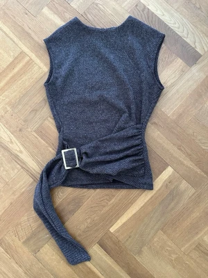 Mörkgrå ärmlös topp med bälte - Snygg mörkgrå ärmlös topp med markerad midja och brett tygbälte med stor metallspänne i sidan. Toppen har en rak halsringning och är gjord i ett mjukt, stickat material som ger en modern och stilren look. Har aldrig använt den, lappen sitter kvar.