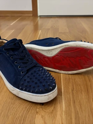 Blå Christian Louboutin sneakers - Snygga mörkblå sneakers från Christian Louboutin med klassiska röda sulor och spikar på tån. Skorna har lågt skaft, snörning och är tillverkade i mocka. Perfekta för dig som vill sticka ut med en exklusiv touch (pris kan diskuteras)