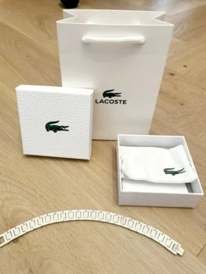 Vitt armband från Lacoste - Snyggt vitt armband från Lacoste i metall med rektangulära länkar och diskret textur på vissa länkar. Armbandet har en stilren och modern design, perfekt för dig som gillar minimalistiska accessoarer. Levereras i originalförpackning med påse och ask.