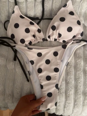 Vit bikini med svarta prickar - Trendig trekantsbikini i vitt med stora svarta prickar och svarta knytband. Klassisk modell med justerbara band både upptill och nedtill. Perfekt för sommarens strandhäng eller poolparty. Osäker på vart den är ifrån då jag fick den i födelsedagspresent och lapparna är avklippta