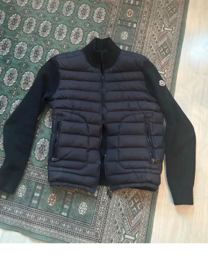 Svart dunväst från Moncler - Snygg svart dunväst från Moncler med quiltad front och två dragkedjefickor. Västen har stickade ärmar och hög krage för extra värme. Klassisk Moncler-logga på ärmen och diskreta detaljer. Perfekt för lager-på-lager och streetstyle.