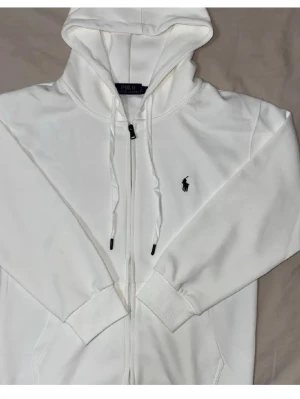 Vit hoodie från Polo Ralph Lauren - Snygg vit hoodie från Polo Ralph Lauren med dragkedja framtill och klassisk svart logga broderad på bröstet. Hoodien har justerbar huva med snören och ribbade muddar vid ärmslut och nederkant. Perfekt för en clean och stilren look.