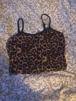 Leopardmönstrad crop top - Snygg leopardmönstrad crop top med tunna svarta axelband och rundad halsringning. Toppen är i stretchigt material och har en tajt passform som framhäver figuren. Perfekt att matcha med höga jeans eller kjol för en trendig look.