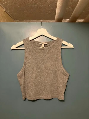 Grå croppad linnetopp från H&M - Säljer en enkel och stilren grå croppad linnetopp från H&M. Toppen är ärmlös med rund halsringning och har en mjuk, ribbad struktur som ger en avslappnad vibe. Perfekt att matcha med höga jeans eller kjol.