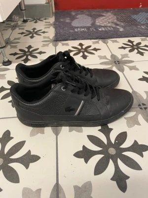 Svarta Lacoste sneakers i läder - Säljer ett par svarta sneakers från Lacoste med klassisk låg profil och snygga detaljer. Skorna är tillverkade i läder med texturerade paneler och har en diskret Lacoste-logga på sidan. Perfekta för dig som gillar stilrena och sportiga skor.