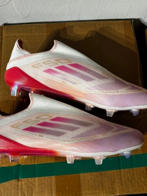 Adidas X Speedflow rosa fotbollsskor - Säljer ett par Adidas X Speedflow fotbollsskor i vitt och rosa med metallicdetaljer. Skorna har en strömlinjeformad design utan snörning, låg vikt och genomskinlig yttersula med dobbar för bästa grepp på planen. Perfekta för dig som vill ha speed och stil på matchen.