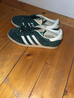 Adidas Gazelle gröna sneakers - Mörkgröna Adidas Gazelle sneakers i mocka med klassiska vita ränder på sidorna och vit hälkappa med grönt Adidas-tryck. Låg modell med gummisula och snörning. Perfekta för en avslappnad streetwear-look.