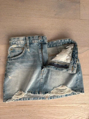 Blå jeanskjol med slitningar - Säljer en kort blå jeanskjol från Pull&Bear med slitna detaljer och rå kant nertill. Kjolen har klassisk femficksdesign, bälteshällor och dragkedja framtill. Perfekt för dig som gillar en avslappnad och trendig stil.