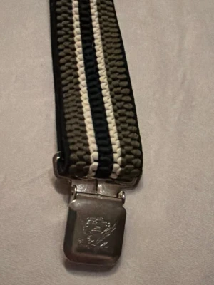 Randiga hängslen med metallclips - Snygga hängslen med breda ränder i olivgrönt, svart och vitt. Hängslena har robusta metallclips med ingraverad detalj framtill. Tillverkade i elastiskt material för skön passform och stilren look.