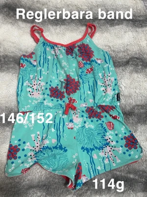 Turkos jumpsuit med havsmotiv - Snygg turkos jumpsuit med justerbara axelband och färgglatt havsmotiv med fiskar, koraller och sjögräs. Röd kant och rosett i midjan ger en extra detalj. Perfekt för sommarens alla äventyr.🌸 Fint använt skick. Reglerbara band. Storlek 146/152. 