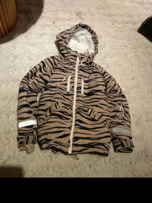 Beige vindjacka med zebramönster - Snygg beige vindjacka från FUNK med svart zebramönster och vita detaljer. Jackan har huva, dragkedja framtill, två stora fickor och reflexband på ärmarna. Perfekt för dig som vill sticka ut med ett coolt mönster och praktiska detaljer.
