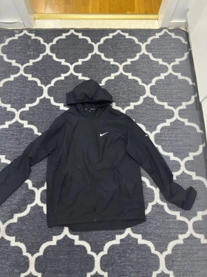 Svart Nike vindjacka med huva - Snygg svart vindjacka från Nike med huva och dragkedja framtill. Jackan har två sidofickor med dragkedja och en vit Nike-logga på bröstet. Tillverkad i lätt och smidigt material, perfekt för löpning eller andra utomhusaktiviteter.