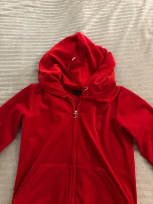 Röd velourhoodie från Juicy Couture - Juicy Couture hoodie i mjuk röd velour med dragkedja framtill och stor huva. Klassisk broderad logga på bröstet och känguruficka. Dragsko i huvan och snygg J-detalj på dragkedjan. Perfekt för en chill och trendig look.
