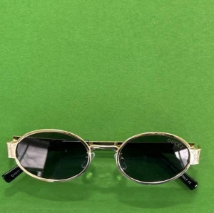 Guldiga ovala solglasögon från Gucci - Snygga solglasögon från Gucci med guldfärgad metallbåge och ovala, mörka linser. Klassisk och stilren design med svarta skalmar och diskret Gucci-logga på linsen. Perfekta för dig som vill ha en exklusiv accessoar med lyxig känsla.