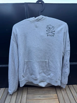 Unik hoodie från Polo Ralph Lauren🙌🤩 - Säljer en grå hoodie  från Polo Ralph Lauren med coolt tryck av björn och korsade ben på bröstet. Tröjan har en klassisk passform, ribbade muddar och en stor magficka. Materialet är mjukt och bekvämt, perfekt för chill dagar. Tryck även på ena ärmen för extra detalj.🙌🤩