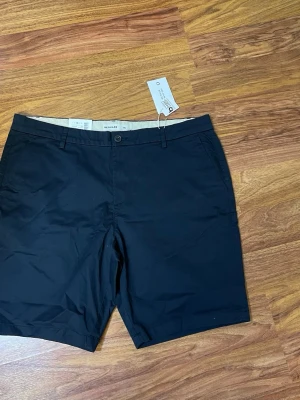 Svarta shorts från Regular - Klassiska svarta shorts från Regular med normal passform. Shortsen har bälteshällor, knapp och dragkedja framtill samt sidofickor och två bakfickor. Perfekta för sommaren och tillverkade i ett mjukt bomullsmaterial.