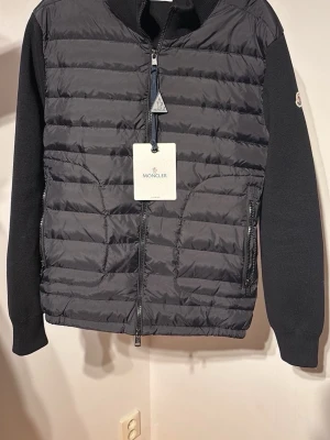Svart dunväst från Moncler - Snygg svart dunväst från Moncler med stickade ärmar och quiltad front. Västen har dragkedja framtill, två sidofickor med dragkedja och Moncler-logga på ärmen. Perfekt för lager-på-lager och riktigt trendig look.