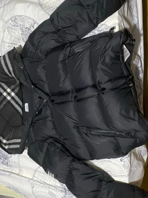 Svart pufferjacka från Burberry - Säljer en svart pufferjacka från Burberry med klassiskt rutigt foder i huvan och tydlig Burberry London England-patch på ärmen. Jackan har dragkedja, tryckknappar och flera fickor. Perfekt för kalla vinterdagar och hösten om du behöver en väst eftersom den kan bli till en väst också pris kan diskuteras