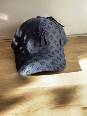 Mörkblå NY keps med mönster - Snygg mörkblå keps med NY-logga framtill och all-over svart mönster. Kepsen har böjd skärm och justerbar passform baktill. Materialet är tyg och ger en trendig streetwear-känsla. Perfekt accessoar för att lyfta din outfit.