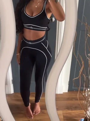Svart träningsset från Alo - Snyggt svart träningsset från Alo med sport-bh och höga tights. Setet har vita kontrastsömmar och logga på både bh och midja. Tillverkat i stretchigt material som sitter tight och bekvämt, perfekt för gymmet eller yoga.