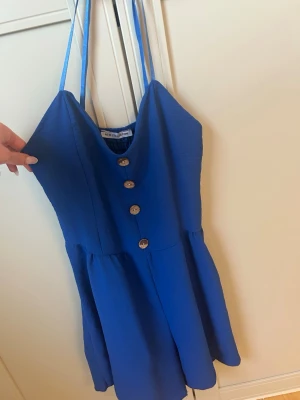 Blå kort shortsdress  med knappar - Snygg blå kort shortsdress med smala axelband och dekorativa knappar framtill. Dressen har en v-ringad urringning och lätt utställd kjol. Perfekt för varma dagar och enkel att styla med accessoarer.