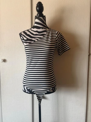 Randig oneshoulder topp i bomull - Snygg svartvit randig topp med oneshoulder-design och kort ärm. Tight passform och ribbad struktur som ger en modern look. Perfekt för dig som vill sticka ut lite extra. Gjord i mjuk bomull för skön känsla.
