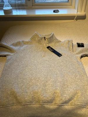 Grå quarterzip  sweatshirt Stone Island - Snygg ljusgrå sweatshirt från Stone Island med halv dragkedja och hög krage. Tröjan har ribbade muddar och den klassiska Stone Island-loggan på ärmen. Perfekt för en avslappnad och stilren look.