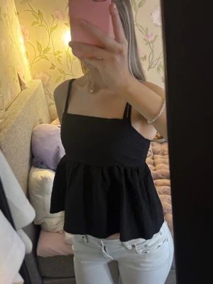 Svart linnetopp med volang - Säljer en svart linnetopp med breda axelband och volang nedtill. Toppen har en rak urringning och sitter löst över magen för en avslappnad look. Perfekt till sommaren och enkel att matcha med olika stilar.