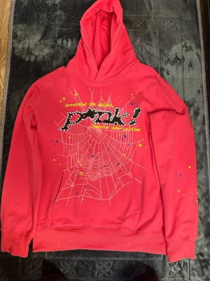 Spider hoodie - Cool röd Spider hoodie med huva och tryck framtill som visar ett spindelnät, texten 'nevermind the spider, forget the slime' och ordet 'prank!' i svartvitt. Små stjärnor i blått och gult pryder både fram- och baksidan, där det även finns en gul stjärna. Perfekt för dig som gillar statement-plagg.
