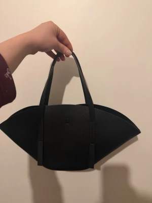 Svart geometrisk handväska - Unik svart handväska i geometrisk form med två långa handtag. Väskan är tillverkad i slätt konstläder och har en minimalistisk design utan synliga loggor. Invändigt finns ett metallspänne för stängning. Perfekt för dig som gillar statement-accessoarer.