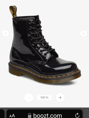 Svarta lackade Dr. Martens boots - Säljer ett par klassiska svarta Dr. Martens boots i glansigt lackmaterial. Skorna har den ikoniska gula sömmen, grov sula och snörning hela vägen upp. Perfekta för dig som vill ha en edgy och tidlös look.