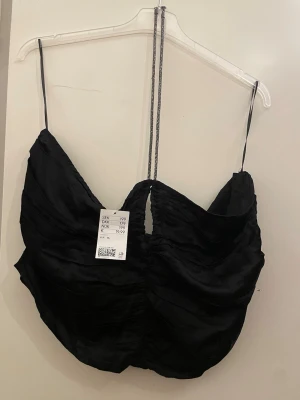 Svart satinlinnetopp med strassband - Snygg svart linnetopp från HM i glansigt satinmaterial. Toppen har tunna axelband med strassdetaljer och en lätt rynkad effekt framtill. Perfekt för dig som vill ha en trendig och elegant look.