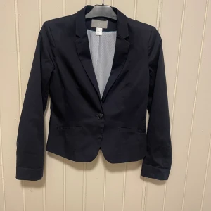 Mörkblå kavaj från H&M, strl 38 - Stilren mörkblå kavaj från H&M i modellen Modern Classic. Kavajen har en knapp framtill, klassisk krage och lätt figursydd passform. Fodret är ljusblått med tunna ränder. Perfekt för en clean och snygg look.