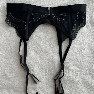 Svart spetsig strumpebandshållare - Säljer en svart strumpebandshållare med vackra spetsdetaljer och justerbara band. Modellen har en bred satinliknande midjerem och metallspännen för att fästa strumpor. Perfekt för att ge din outfit en extra touch av elegans.