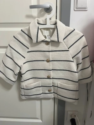 Vit randig overshirt med knappar - Stilren vit overshirt med svarta horisontella ränder och träknappar framtill. Jackan har två stora fickor fram, krage och en boxig, croppad passform med halvlånga ärmar. Perfekt för lager på lager och en avslappnad look.