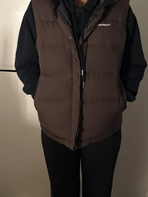 Brun dunväst från Everest - Mörkbrun dunväst från Everest med hög krage och dragkedja framtill. Västen har en puffig, quiltad design och en stor huva som ger extra värme. Perfekt för lager-på-lager och funkar till många olika stilar.