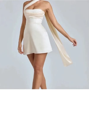 Klänning - Snygg miniklänning från OhPolly i champagnefärg med oneshoulder-design och lång scarfdetalj. Klänningen har snörning i ryggen och är tillverkad i ett glansigt material som ger en lyxig känsla. Perfekt för dig som vill sticka ut med en unik och trendig look. Använd endast en kväll.