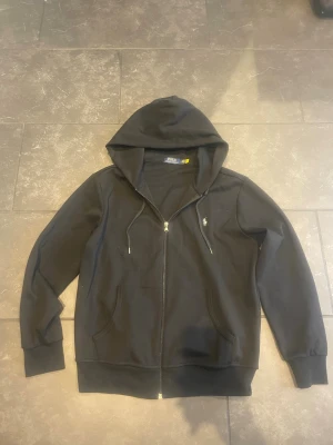 Svart hoodie från Polo Ralph Lauren - Snygg svart hoodie med dragkedja från Polo Ralph Lauren. Klassisk design med huva och dragsnören, två fickor framtill och den ikoniska broderade loggan på bröstet. Perfekt för en chill och stilren look.