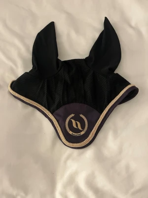 Svart huva från Back on Track - Säljer en svart huva från Back on Track med guldigt kantband och broderad logga framtill. Tillverkad i meshmaterial som andas och har två långa öron. Perfekt för dig som vill ha en snygg och funktionell accessoar till hästen.