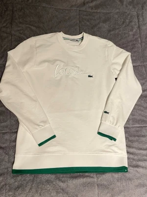 Vit långärmad topp från Lacoste - Säljer en vit långärmad topp från Lacoste med broderad logga och krokodil på bröstet. Tröjan har gröna detaljer vid ärmslut och nederkant samt rund halsringning. Perfekt för dig som gillar klassisk och sportig stil.