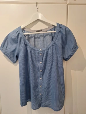 Blå randig blus från Marc O'Polo - Snygg blå blus från Marc O'Polo med tunna vita ränder och rundad halsringning. Blusen har korta ärmar, knappar framtill och en liten knytning mitt bak. Tillverkad i ett lätt och luftigt material som passar perfekt till sommaren. Enkel och stilren design.