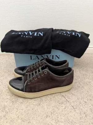 Lanvin bruna sneakers med lackdetalj - Snygga bruna sneakers från Lanvin med ovandel i mocka och svart lackad tå. Skorna har beige sula och klassisk låg siluett med svarta skosnören. Perfekta för dig som gillar stilrena och exklusiva sneakers med unika materialval.