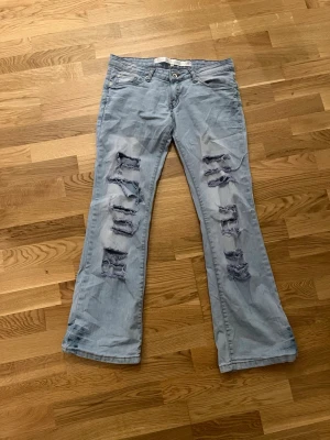 Ljusblå slitna bootcut jeans - Säljer ett par ljusblå bootcut jeans med flera slitningar och hål framtill. Jeansen har fem fickor, låg midja och broderade detaljer på bakfickorna. Perfekta för en avslappnad och trendig look.