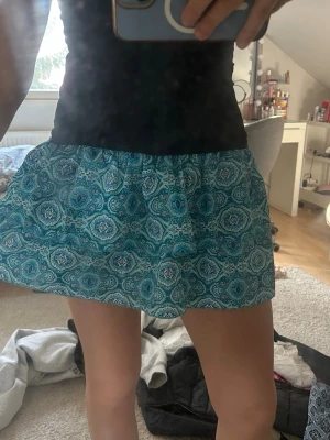 Blå mönstrad kortkjol - Kortkjol i blått med ett detaljerat mönster i vitt och turkos. Kjolen har en avslappnad passform med resår i midjan och är tillverkad i ett lätt och luftigt material, perfekt för varma dagar.