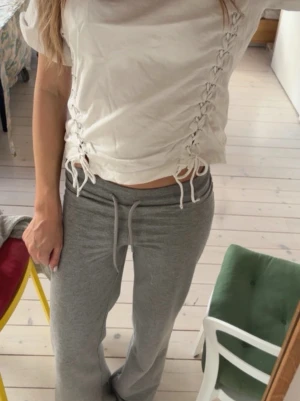 Vit topp med snörning i sidorna - Trendig vit topp med snörning längs båda sidorna som ger en cool och unik look. Modellen är croppad och har korta ärmar. Perfekt för dig som vill ha en enkel men ändå detaljerad topp. Tillverkad i mjukt bomullsmaterial.