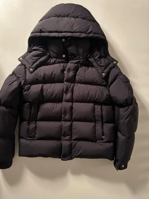 Moncler Vezere - Snygg svart pufferjacka med stor vadderad huva och dragkedja samt tryckknappar framtill. Jackan har två sidofickor med dragkedja och breda quiltade paneler för extra värme. Perfekt för kalla vinterdagar och riktigt skön att bära.