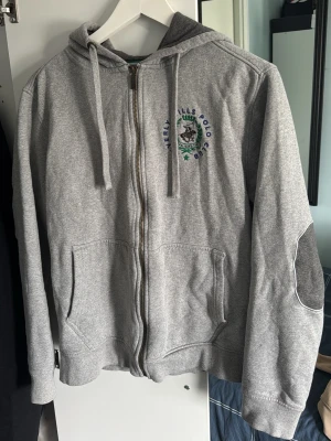 Grå zip-hoodie Beverly Hills Polo Club - Snygg grå zip-hoodie från Beverly Hills Polo Club med broderad logga på bröstet och kontrasterande armbågslappar. Hoodien har dragsko i huvan, två fickor fram och ribbade muddar. Perfekt för en avslappnad och sportig stil. Köptes utomlands, finns ej i Sverige