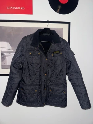 Svart quiltad rock från Barbour - Stilren svart quiltad rock från Barbour med klassisk krage i manchestertyg, tryckknappar i mässing och flera fickor framtill. Rocken har en tidlös design och Barbour-logga på bröstet. Perfekt för dig som gillar brittisk stil. Obs jackan har bara knappar. Ingen dragkedja. Vid snabb affär kan jag gå ner en hundring.