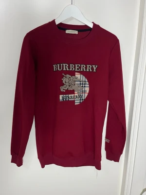 Vinröd sweatshirt från Burberry - Vinröd sweatshirt från Burberry med klassiskt tryck och logga på bröstet samt rutigt mönster. Rund halsringning och långa ärmar. Perfekt för dig som gillar streetwear och exklusiva märken.